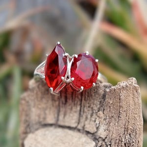 Può includere: Un anello d'argento con due grandi pietre preziose rosse. L'anello è posto su un pezzo di legno.