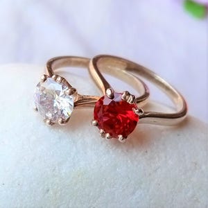 Könnte beinhalten: Zwei goldene Ringe mit klaren und roten Edelsteinen. Die Ringe liegen auf einer weißen Oberfläche.