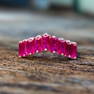 May include: A silver ring with a row of bright fuchsia-coloured, rectangular gemstones. The gemstones are set close together, forming a continuous band of colour. The ring is displayed on a textured wooden surface.
