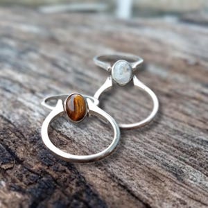 Könnte beinhalten: Zwei silberne Ringe mit ovalen Edelsteinen. Der Ring links hat einen braunen Tigerauge-Stein. Der Ring rechts hat einen weißen Mondstein.