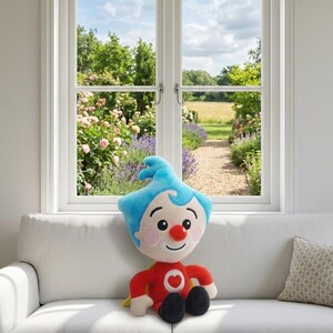 Op de afbeelding: Een pluche pop met een vrolijk gezicht, een rood shirt met een hart, blauw haar en zwarte schoenen. De pop zit op een witte bank voor een raam met uitzicht op een tuin.