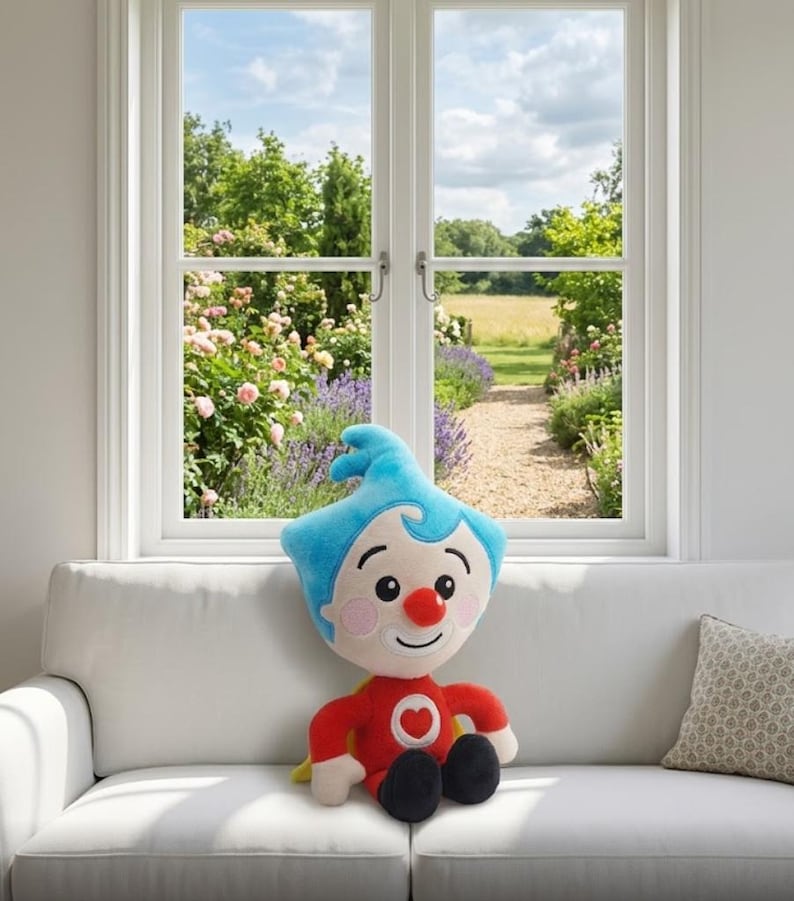 Op de afbeelding: Een pluche pop met een vrolijk gezicht, een rood shirt met een hart, blauw haar en zwarte schoenen. De pop zit op een witte bank voor een raam met uitzicht op een tuin.