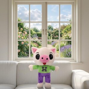 Op de afbeelding: Een pluchen varken met een lichtroze gezicht, oren en snuit, en grote zwarte ogen. Het draagt een groen shirt met een zwarte "H" en een paarse broek. Het speelgoed staat op een witte bank.