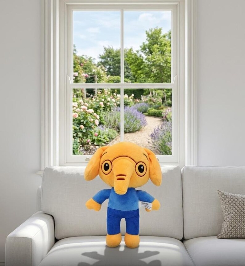 Op de afbeelding: Een pluche olifant met een gele kop, oren en voeten, en een blauw shirt en broek. Het speelgoed heeft grote, ronde ogen en een slurf. Het staat op een witte bank voor een raam met uitzicht op een tuin.