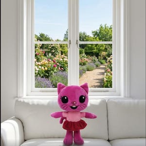 Op de afbeelding: Een roze pluche kattenpop met grote zwarte ogen, een lachend gezicht en een rode rok. De pop staat op een witte bank voor een raam met uitzicht op een bloementuin.