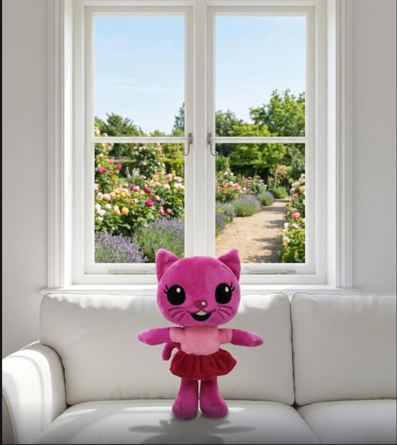Op de afbeelding: Een roze pluche kattenpop met grote zwarte ogen, een lachend gezicht en een rode rok. De pop staat op een witte bank voor een raam met uitzicht op een bloementuin.