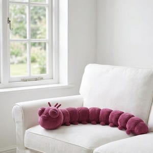 Pode incluir: Uma almofada de pelúcia em forma de lagarta bordô com cabeça arredondada, olhos grandes e corpo segmentado. O brinquedo macio está em um sofá branco perto de uma janela, oferecendo um elemento decorativo lúdico.
