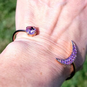 Könnte beinhalten: Ein goldfarbenes Armreif-Armband mit einem großen, quadratischen, lila Edelstein und einem Mond-Design. Der Mond ist mit kleineren lila Kristallen besetzt. Das Armband ist offen und wird am Handgelenk getragen.