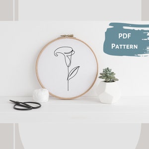 May include: A white embroidery hoop with a black line drawing of a calla lily. The hoop is on a white surface with a small potted succulent and a pair of scissors.