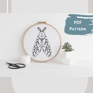 May include: A black and white embroidery pattern of a moth with intricate details on its wings. The pattern is displayed in a wooden embroidery hoop on a white surface. The text "PDF PATTERN" is written in blue on a gray background.