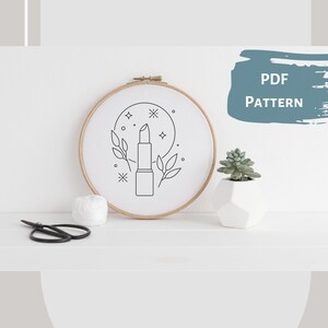 Pode incluir: Um desenho em preto e branco de um batom com folhas e estrelas em um círculo em um tecido branco em um aro de bordado de madeira. O aro está sobre uma superfície branca com uma pequena planta suculenta em vaso e um par de tesouras pretas.