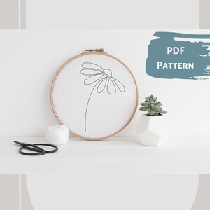 May include: A simple line drawing of a daisy flower in a wooden embroidery hoop. The hoop is on a white surface with a small potted succulent plant and a pair of scissors.