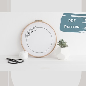 May include: A black and white embroidery pattern of a simple floral wreath with a circle in the center. The pattern is displayed in a wooden embroidery hoop on a white surface. The text "PDF PATTERN" is written in blue on a white background.