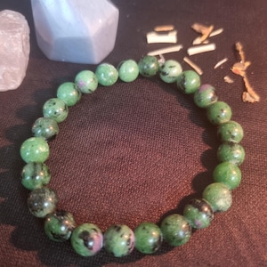 May include: A beaded bracelet composed of round, polished stones in shades of green, black, and occasional flecks of magenta. The beads are strung together in a circular loop, showcasing natural variations in colour and texture throughout the piece.