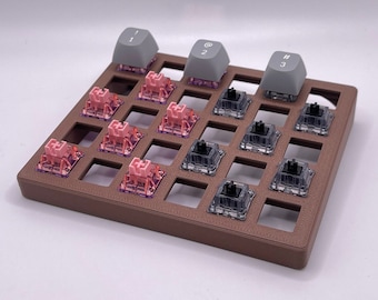 May include: A brown plastic keyboard keycap holder with 16 slots. The holder is filled with various colored keycaps, including pink, gray, and black. The keycaps have different symbols printed on them, including numbers, letters, and punctuation marks.
