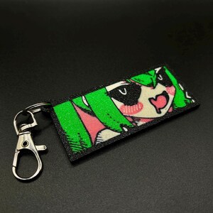Puede incluir: Un llavero rectangular con un personaje de estilo anime con cabello verde, mejillas rosadas y boca en forma de coraz&oacute;n. El llavero tiene un borde negro y un cierre plateado.