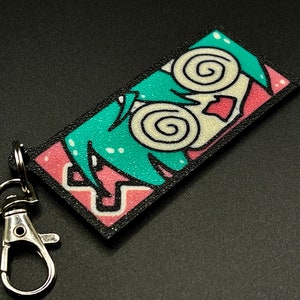 May include: A black and pink keychain with a cartoon character with green hair and swirling eyes. The character has a pink mouth and a white face.