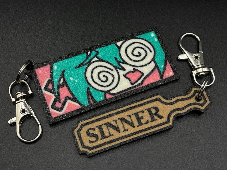 May include: A black and white keychain with a colorful anime character with green hair and the word "SINNER" on a brown tag.