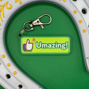 Peut inclure: Un porte-clés en forme de fer à cheval blanc avec des détails dorés et une étiquette de clé verte avec le texte "Umazing!" et un symbole de pouce levé. Le porte-clés est argenté et le fond est vert.
