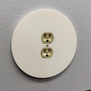 May include: A white, circular electrical outlet cover with two standard outlets. The outlets are beige and set against a light gray wall. The cover is flush with the wall.