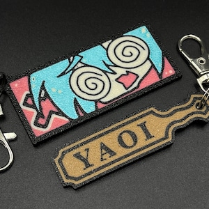 May include: A blue and pink anime-style keychain with a swirling eye design and the word "YAOI" on a brown tag.