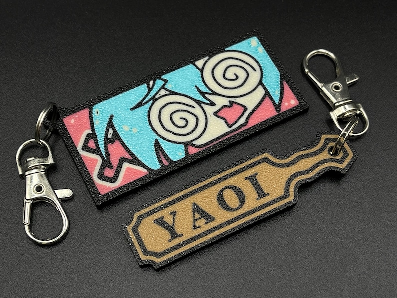 May include: A blue and pink anime-style keychain with a swirling eye design and the word "YAOI" on a brown tag.