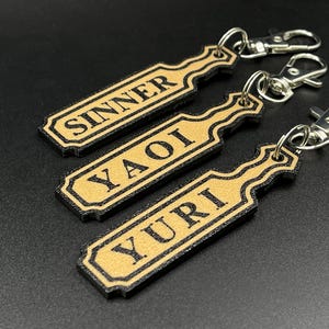 May include: Three gold-colored keychains with black trim and silver key rings. The keychains are shaped like paddles and have the words "Sinner", "Yaoi", and "Yuri" printed on them.