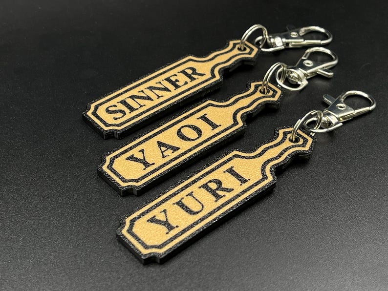May include: Three gold-colored keychains with black trim and silver key rings. The keychains are shaped like paddles and have the words "Sinner", "Yaoi", and "Yuri" printed on them.