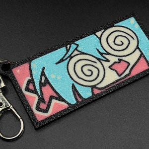 May include: A black and white cartoon character with blue hair and pink and white background on a black keychain.