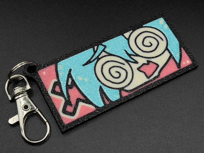 May include: A black and white cartoon character with blue hair and pink and white background on a black keychain.