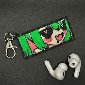 Puede incluir: Un llavero rectangular con un dise&ntilde;o de personaje de anime, con cabello verde, mejillas rosadas y una boca en forma de coraz&oacute;n. El llavero tiene un cierre plateado y est&aacute; junto a un par de auriculares inal&aacute;mbricos blancos.