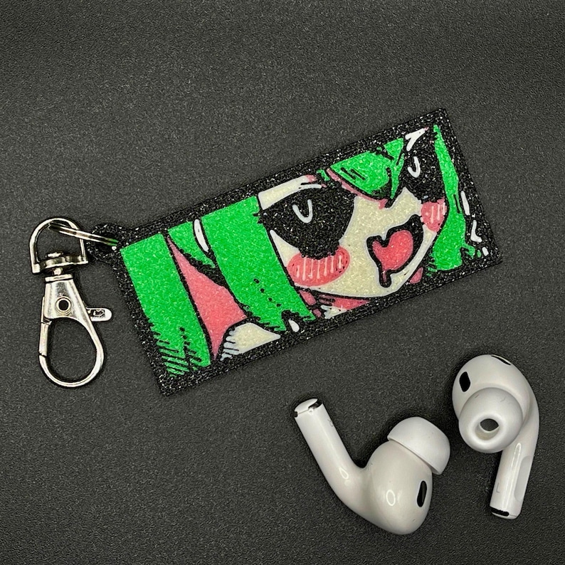 Puede incluir: Un llavero rectangular con un dise&ntilde;o de personaje de anime, con cabello verde, mejillas rosadas y una boca en forma de coraz&oacute;n. El llavero tiene un cierre plateado y est&aacute; junto a un par de auriculares inal&aacute;mbricos blancos.
