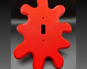 May include: A bright red, irregularly shaped light switch cover with a single switch opening. The cover is designed to look like a splatter of paint.