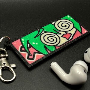 May include: A black and pink keychain with a cartoon character with green hair and swirling eyes. The keychain is attached to a silver metal key ring.