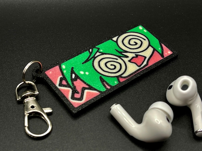 May include: A black and pink keychain with a cartoon character with green hair and swirling eyes. The keychain is attached to a silver metal key ring.