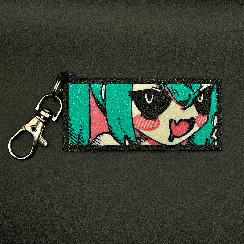 Puede incluir: Un llavero rectangular con la cara de un personaje de estilo anime colorido con pelo verde azulado, mejillas rosadas y boca roja. El llavero tiene un cierre plateado y un borde negro.