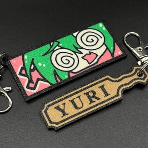 May include: A black and pink keychain with a cartoon character with green hair and swirling eyes. The keychain also includes a gold tag with the word "YURI" printed on it.