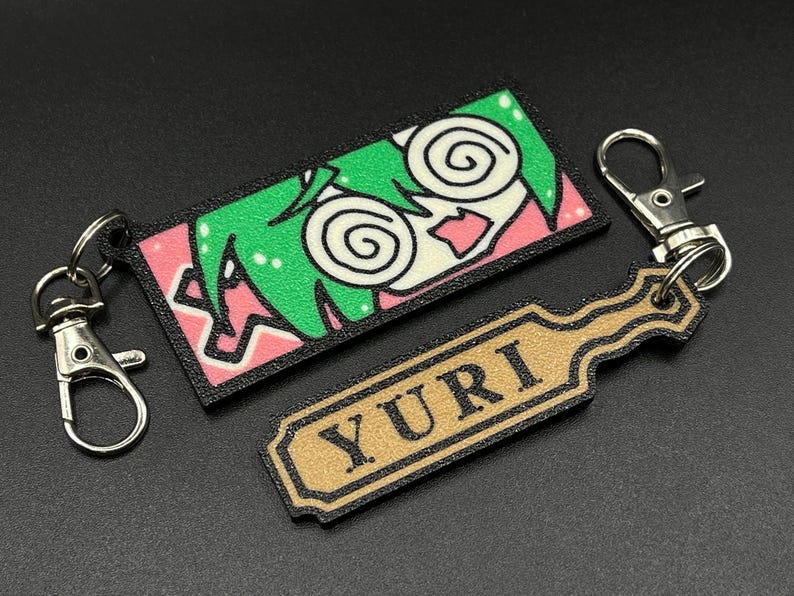 May include: A black and pink keychain with a cartoon character with green hair and swirling eyes. The keychain also includes a gold tag with the word "YURI" printed on it.