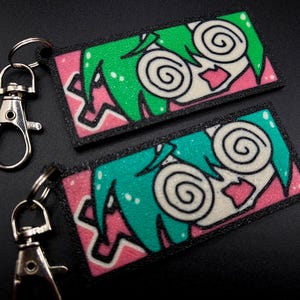 May include: Two keychains with black and white cartoon faces with green and blue hair. The faces have pink lips and large, swirling eyes. The keychains are on a black surface.
