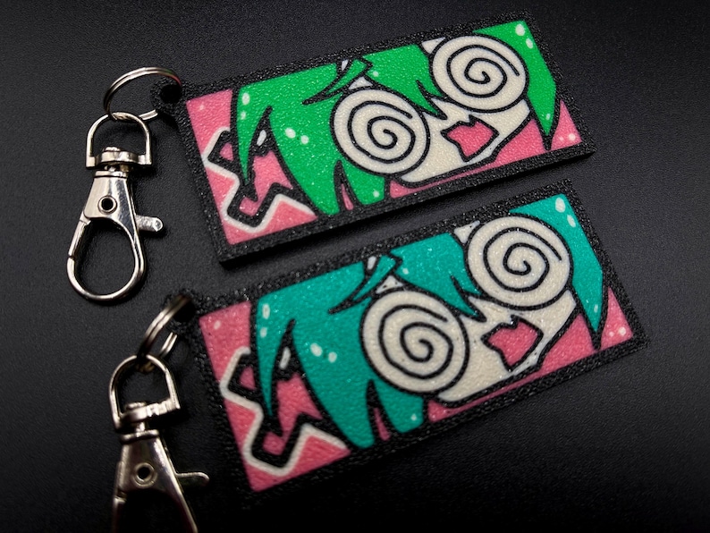 May include: Two keychains with black and white cartoon faces with green and blue hair. The faces have pink lips and large, swirling eyes. The keychains are on a black surface.