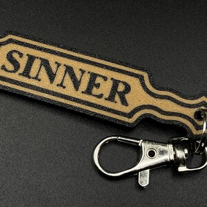 May include: A black and gold key chain with a paddle-shaped charm that says "SINNER" in black lettering.