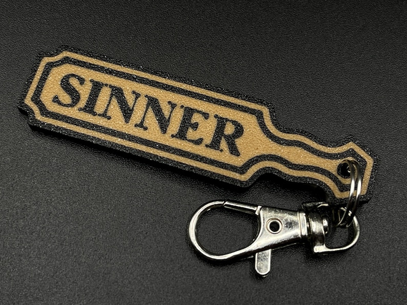 May include: A black and gold key chain with a paddle-shaped charm that says "SINNER" in black lettering.
