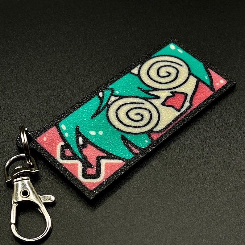 May include: A black and pink keychain with a cartoon character with green hair and swirling eyes. The character has a pink mouth and a white face.