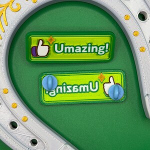 May include: A white horseshoe with gold accents and two green rectangular magnets. The magnets have the text "Umazing!" and "gnisemu" with a thumbs-up icon and a blue sphere. The background is green.