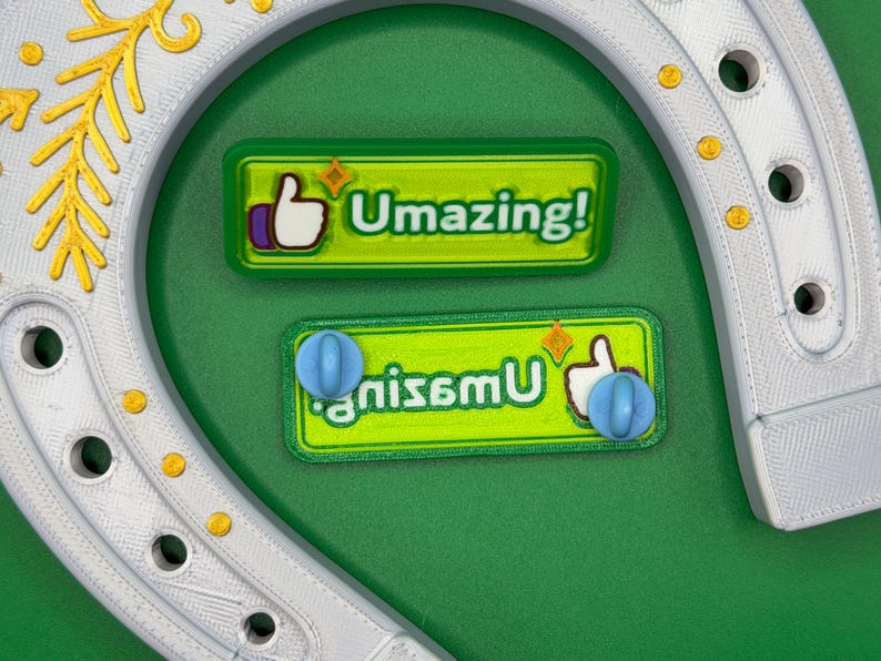 May include: A white horseshoe with gold accents and two green rectangular magnets. The magnets have the text "Umazing!" and "gnisemu" with a thumbs-up icon and a blue sphere. The background is green.