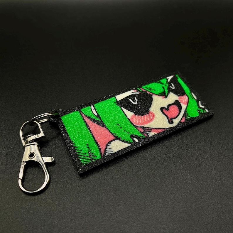 Puede incluir: Un llavero rectangular con un personaje de estilo anime con cabello verde, mejillas rosadas y boca en forma de coraz&oacute;n. El llavero tiene un borde negro y un cierre plateado.