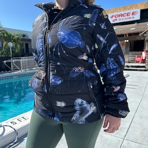 Könnte beinhalten: Schwarze Steppjacke mit einem Muster aus blauen und weißen Meereskreaturen. Die Jacke hat einen hohen Kragen und einen Reißverschluss. Die Ärmel sind lang und die Jacke ist gesteppt. Die Person trägt grüne Leggings. Im Hintergrund ein Pool und ein Schild mit der Aufschrift "FORCE E SCUBA".