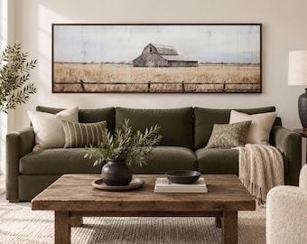 May include: A rectangular wall art print featuring a weathered barn in a golden field, framed in dark wood. The scene depicts a rustic landscape with a wooden fence in the foreground, displayed above a dark olive green sofa with various throw pillows.