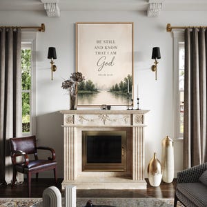 May include: A framed artwork with the text "BE STILL AND KNOW THAT I AM God" above a landscape painting of a river and trees. The artwork hangs above a fireplace with decorative vases and candles. The room has a classic, elegant design.