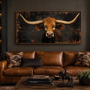 May include: A rectangular wall art piece featuring a longhorn cow portrait with large curved horns against a dark, textured background. The artwork is framed in a thin brown border and is centered above a brown leather sofa with patterned pillows.